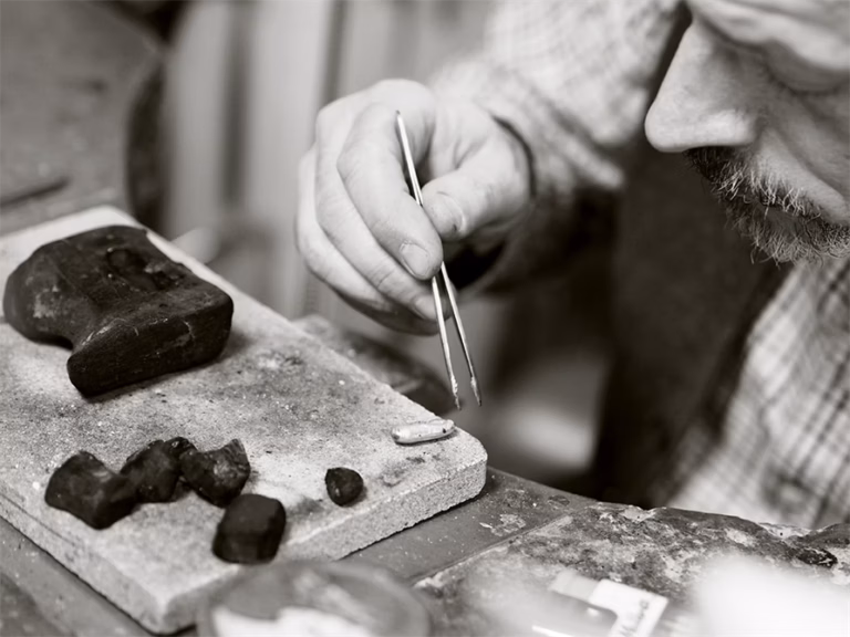 Svartvit bild på en av våra guldsmeder som håller i en tång för att ta i en varm silverbit som ska bli till en ring.